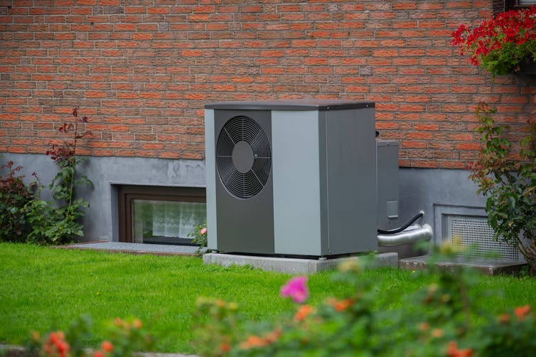 Gray air source heat pump unit on exterior of old red brick house, green grass and flowers in foreground